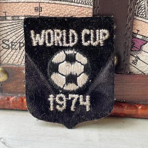 May include: A black shield-shaped embroidered patch featuring the white text WORLD CUP at the top and the year 1974 at the bottom. In the centre, there is a white and black stitched football graphic. The patch has a textured fabric surface.
