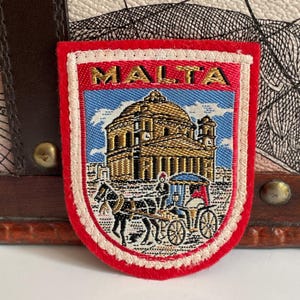 Op de afbeelding: Een schildvormige geborduurde patch met het woord MALTA in gouden letters aan de bovenkant. Het ontwerp toont een groot gebouw met koepel in goud- en bruintinten boven een zwarte paardenkoets, alles tegen een lichtblauwe luchtachtergrond.