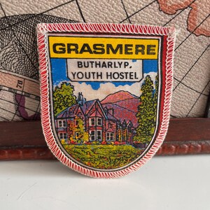 Könnte beinhalten: Ein gestickter Aufnäher in Rot, Weiß und Blau mit dem Text "Grasmere Buttarlyp. Youth Hostel". Der Aufnäher zeigt ein Cartoon-Bild eines Gebäudes mit einem Berg im Hintergrund.