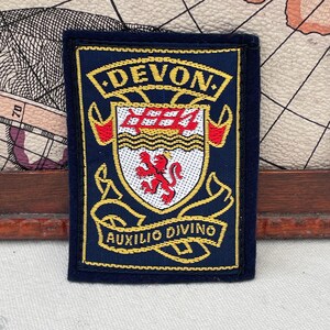 May include: A navy blue embroidered patch with gold lettering and a crest. The text "DEVON" is at the top, and "AUXILIO DIVINO" is at the bottom. The crest features a red lion and a red and gold design.