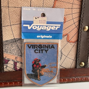 May include: A vintage Voyager originals patch featuring a design of Virginia City. The patch is blue with an embroidered image of a person in a red hat and coat. The patch is in a clear plastic sleeve with a price tag.