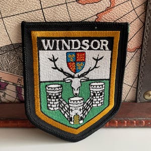 Könnte beinhalten: Gestickter Aufnäher mit dem Wort "WINDSOR" oben. Der Aufnäher zeigt einen Hirschkopf mit Geweih, ein Schild und Burgillustrationen in Grün, Weiß und Gold. Der Aufnäher hat einen schwarzen Rand.
