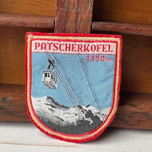 May include: A vintage patch with a red border and the text "PATSCHERKOFEL 2250m". The patch features a blue background with a cable car graphic and a mountain range at the bottom. The patch is rectangular with rounded corners.