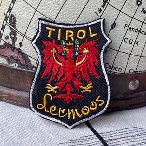 Könnte beinhalten: Ein schwarz-weißer, gestickter Aufnäher mit dem Wort "TIROL" oben und "Lermoos" unten in goldfarbenen Buchstaben. Der Aufnäher zeigt ein rotes Adleremblem mit goldener Krone und Olivenzweigen.