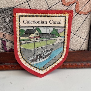 以下が含まれることがあります： 上部に「Caledonian Canal」の文字が入った赤とクリーム色のパッチ。パッチには、ボート、建物、木々のある運河のイラストが描かれています。パッチは黄色のステッチで縁取られています。