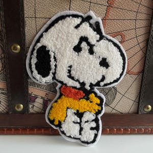 Könnte beinhalten: Gestickter Aufnäher mit Snoopy aus dem Comic Peanuts. Der Aufnäher hat einen weißen Körper, schwarze Ohren und Augen sowie einen gelb-orangen Schal. Der Aufnäher befindet sich auf einem Hintergrund mit einem Vintage-Kartenmuster.