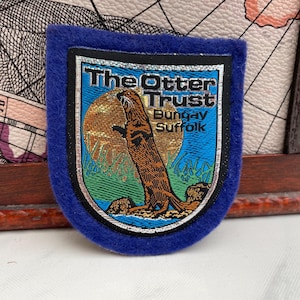 Könnte beinhalten: Blauer Aufnäher mit dem Text "The Otter Trust Bungay Suffolk". Der Aufnäher zeigt eine Otter-Illustration vor einer goldenen Sonne, mit grünem Gras und blauem Wasser. Der Aufnäher hat einen blauen Rand.
