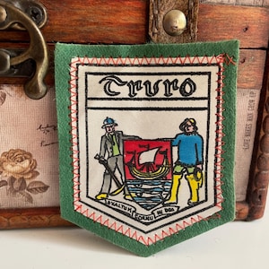 May include: A green felt patch with a white center featuring the word "Truro" in a decorative font. The patch has a crest design with figures, a ship, and text. Red stitching outlines the white center and the green border.