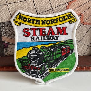 May include: Embroidered patch with a steam train design, featuring the text "North Norfolk Steam Railway" in red and yellow. The patch has a white border and the town name "Sheringham" at the bottom, a souvenir of the area.