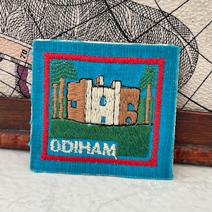 May include: A square, embroidered patch with a teal background and a red border. The patch features a detailed depiction of a building with brown, white, and tan sections, set against a green grassy area. The word "ODIHAM" is embroidered at the bottom.