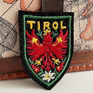 Könnte beinhalten: Ein schwarzer, gestickter Aufnäher mit dem Wort "TIROL" in gelben Buchstaben. Der Aufnäher zeigt ein rotes Adleremblem mit gelben Akzenten und einem grünen Rand. Am unteren Rand des Aufnähers befindet sich eine weiße und gelbe Blume.