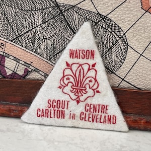 May include: A white triangular Scout patch with red text and a fleur-de-lis emblem. The text reads "WATSON" at the top, with "SCOUT CENTRE CARLTON in CLEVELAND" below. The patch is likely vintage.