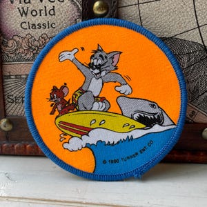 May include: A circular embroidered patch with a bright orange background and a blue border. It features Tom the cat surfing on a yellow surfboard, with Jerry the mouse nearby and a shark in the water. The text 1990 TURNER ENT. CO. is printed.
