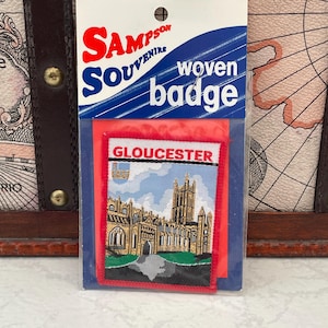 May include: A woven badge souvenir from Sampson Souvenirs, depicting Gloucester Cathedral. The badge, with a red border, features the word "GLOUCESTER". The packaging is blue and white, with the words "woven badge".
