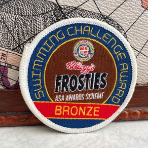 May include: A round, embroidered patch with the text "SWIMMING CHALLENGE AWARD" in blue. The center features "Kellogg's FROSTIES" in white, with "ASA AWARDS SCHEME" below and "BRONZE" in red.