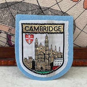 Op de afbeelding: Een lichtblauwe vilten patch met een geborduurd wapenschild en het woord CAMBRIDGE in gouden letters. Het ontwerp bevat een gedetailleerde afbeelding van een gotisch gebouw in gouden en zwarte draad, naast een klein rood-wit schildembleem.