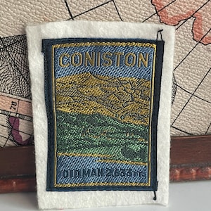 Puede incluir: Un parche rectangular bordado que muestra un paisaje de montaña en tonos dorados y verdes. El texto CONISTON está bordado en oro en la parte superior, y OLD MAN 2.633 FT está bordado en oro en la parte inferior sobre un fondo azul y fieltro.