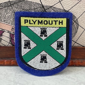 May include: A shield-shaped embroidered patch with a blue felt border. The center features a white field divided by a green saltire, with four black castle icons positioned in the quadrants. The word PLYMOUTH is stitched in black on a yellow band.