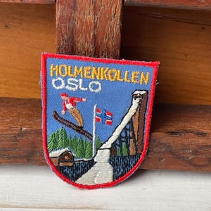 May include: Embroidered patch with the text "HOLMENKOLLEN OSLO" above a skier, ski jump, Norwegian flag, and trees. The patch has a blue background with a red border.