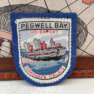Op de afbeelding: Een blauwe patch met witte stiksels. Het bevat de woorden "PEGWELL BAY HOVERPORT" en "RAMSGATE CALAIS". De patch toont een hovercraft met rode en grijze details, varend op blauw water.