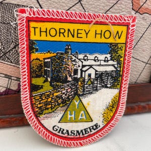 May include: A vintage patch with a red border and white stitching. The patch features the text "THORNEY HOW" and "GRASMERE" above and below a colorful illustration of a building and landscape. A yellow triangle with "Y" and "HA" is also present.