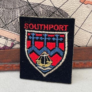 Puede incluir: Un parche rectangular negro con la palabra "SOUTHPORT" en letras rojas en la parte superior. El parche presenta un diseño de escudo con detalles bordados en rojo, azul y amarillo. El escudo tiene un patrón azul ondulado y una estructura amarilla.