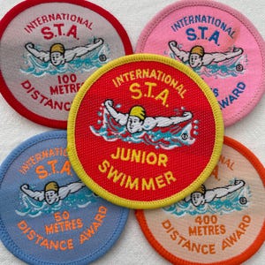 Pode incluir: Uma coleção de cinco patches bordados circulares. Cada patch apresenta o texto "INTERNATIONAL S.T.A." e um gráfico de nadador. Os patches são em várias cores, incluindo vermelho, rosa, azul e laranja, com texto indicando distâncias em metros.
