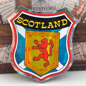 Könnte beinhalten: Ein blauer, weißer und roter schildförmiger Aufkleber mit dem Wort "SCOTLAND" in gelben Buchstaben. Der Schild zeigt einen goldenen Löwen rampant auf rotem Hintergrund.