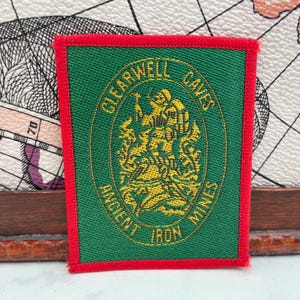 Könnte beinhalten: Grüner und roter Aufnäher mit dem Text "CLEARWELL CAVES ANCIENT IRON MINES" in goldener Schrift. Der Aufnäher zeigt eine goldene Illustration einer Figur mit einer Spitzhacke.