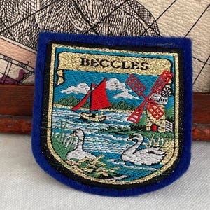 Può includere: Toppa ricamata con il nome "BECCLES" in lettere dorate. Raffigura una barca a vela rossa, un mulino a vento e due cigni su sfondo blu, con un bordo dorato e un bordo in feltro blu.