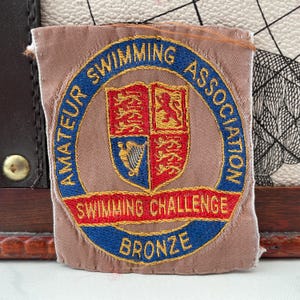 May include: A rectangular patch featuring the text "AMATEUR SWIMMING ASSOCIATION" in blue, encircling a shield with a red and blue emblem. The text "SWIMMING CHALLENGE" is in red, and "BRONZE" is in gold.