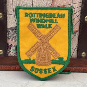 May include: A shield-shaped embroidered patch with a yellow background and green border. It features a brown windmill graphic in the centre, with the green text ROTTINGDEAN WINDMILL WALK above and the green text SUSSEX at the bottom of the patch.