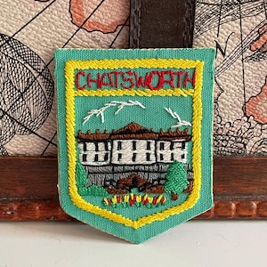 Op de afbeelding: Een schildvormige geborduurde patch met een lichtgroene achtergrond en een gele rand. De tekst CHATSWORTH is bovenaan in het rood geborduurd. Daaronder staat een wit gebouw met bruine dakdetails boven kleurrijke bloemaccenten.