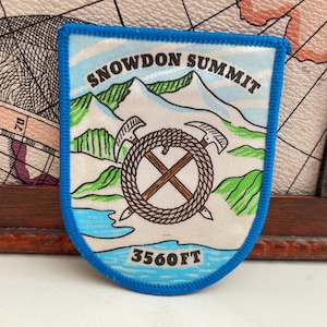 Może przedstawiać: Niebieska naszywka z napisem "SNOWDON SUMMIT" nad górskim krajobrazem. Naszywka przedstawia okrąg z liny ze skrzyżowanymi toporami i napisem "3560FT" na dole, wskazującym wysokość szczytu w stopach (około 1085 metrów).