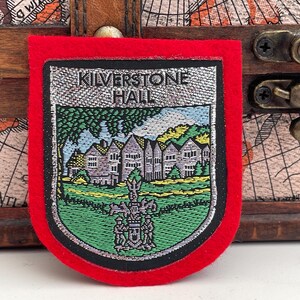 Könnte beinhalten: Ein roter Filz-Patch mit einem silbernen gestickten Bild eines Gebäudes mit dem Text "Kilverstone Hall" darüber.