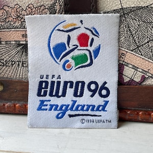 May include: A rectangular white fabric patch featuring the UEFA Euro 96 England logo. The design includes a stylised football graphic with blue, red, green, and yellow accents, the text UEFA EURO 96 England, and the copyright text 1994 UEFA TM.