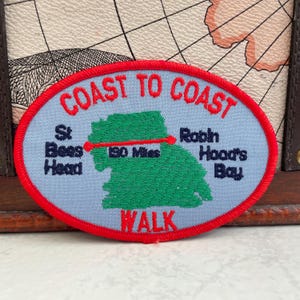 May include: An oval patch with a red border and the words "COAST TO COAST" in red. The patch features a light blue background with a green map and the text "St Bees Head" and "Robin Hood's Bay". A red line indicates a 306 km walk.