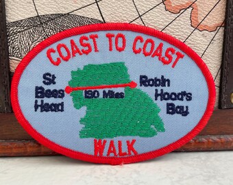 Op de afbeelding: Ovale patch met een rode rand en de rode woorden "COAST TO COAST". De patch heeft een lichtblauwe achtergrond met een groene kaart en de tekst "St Bees Head" en "Robin Hood's Bay". Een rode lijn geeft een wandeling van 306 km aan.