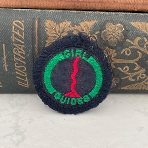 Puede incluir: Una insignia redonda de color azul oscuro de las Girl Guides con un círculo verde y una línea roja ondulada en el centro. El texto "Girl Guides" está bordado en blanco.
