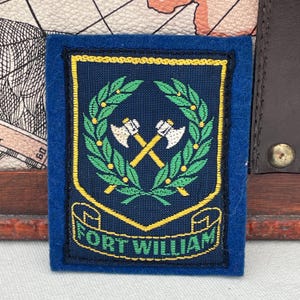 May include: A navy blue patch with a gold border and the text "FORT WILLIAM". The patch features a crossed axe design within a green laurel wreath. The patch is rectangular with a blue border.