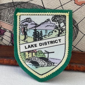 Op de afbeelding: Een schildvormige stoffen embleem met een groene rand en gele stiksels. Het midden toont een bedrukt landschap van bergen, een meer, bomen en een stenen brug. De tekst LAKE DISTRICT is in het zwart gedrukt op een witte banner.