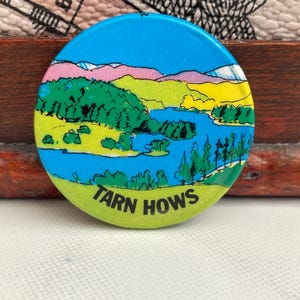 May include: A round button with a landscape design featuring a lake, trees, and mountains under a blue sky. The text "TARN HOWS" is printed in white on a green background at the bottom of the button.