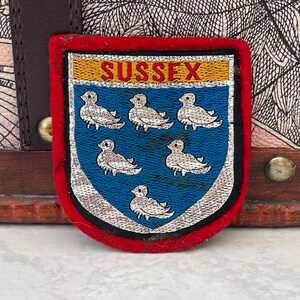 May include: A shield-shaped patch with a red border, featuring a blue center with six embroidered white birds. The word "SUSSEX" is embroidered in gold across the top. The patch is likely a vintage emblem or crest.