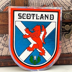May include: A red, white and blue sticker with the word "SCOTLAND" in a stylised font. The sticker features a red lion rampant on a white cross against a blue background. A green thistle is at the bottom of the sticker.