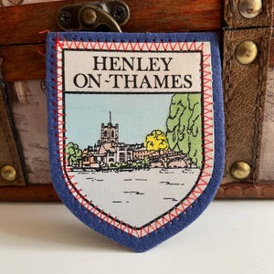 Peut inclure: Écusson bleu en forme de bouclier avec le texte "HENLEY ON-THAMES" et une illustration colorée d'un paysage urbain. Le patch a des coutures rouges, probablement un souvenir ou un emblème de la ville.