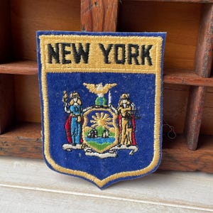 Puede incluir: Parche bordado azul y dorado con las palabras "NEW YORK" en la parte superior. El parche tiene un diseño de escudo con el escudo de armas del estado de Nueva York en el centro. El parche es rectangular con la parte inferior puntiaguda.