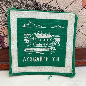 Pode incluir: Remendo quadrado verde com uma borda branca. O remendo apresenta uma ilustração branca de um edifício, nuvens e uma cerca. O texto "AYSGARTH Y H" é impresso em branco na parte inferior do remendo.