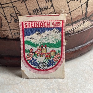 Könnte beinhalten: Ein weißer Stoff-Patch mit roten, blauen und grünen Stickereien. Der Patch zeigt eine Berglandschaft mit einem kleinen Dorf und dem Text "STEINACH O.B. TIROL".