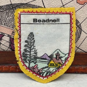 May include: A shield-shaped fabric patch with a yellow border and red zigzag stitching. The centre features a white background with the text Beadnell printed in black. Below the text is a landscape illustration of a tall tree and a yellow house.