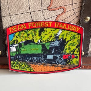 Könnte beinhalten: Rechteckiger Aufnäher mit einem grünen Dampfzug und dem Text "DEAN FOREST RAILWAY" in Rot. Der Zug fährt auf einer Strecke mit grünem und gelbem Hintergrund. Der Aufnäher hat einen roten Rand.